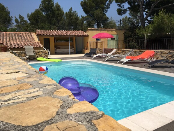 En swimmingpool med stenplattor, solstolar och en liten byggnad med ett tak täckt av plattor.