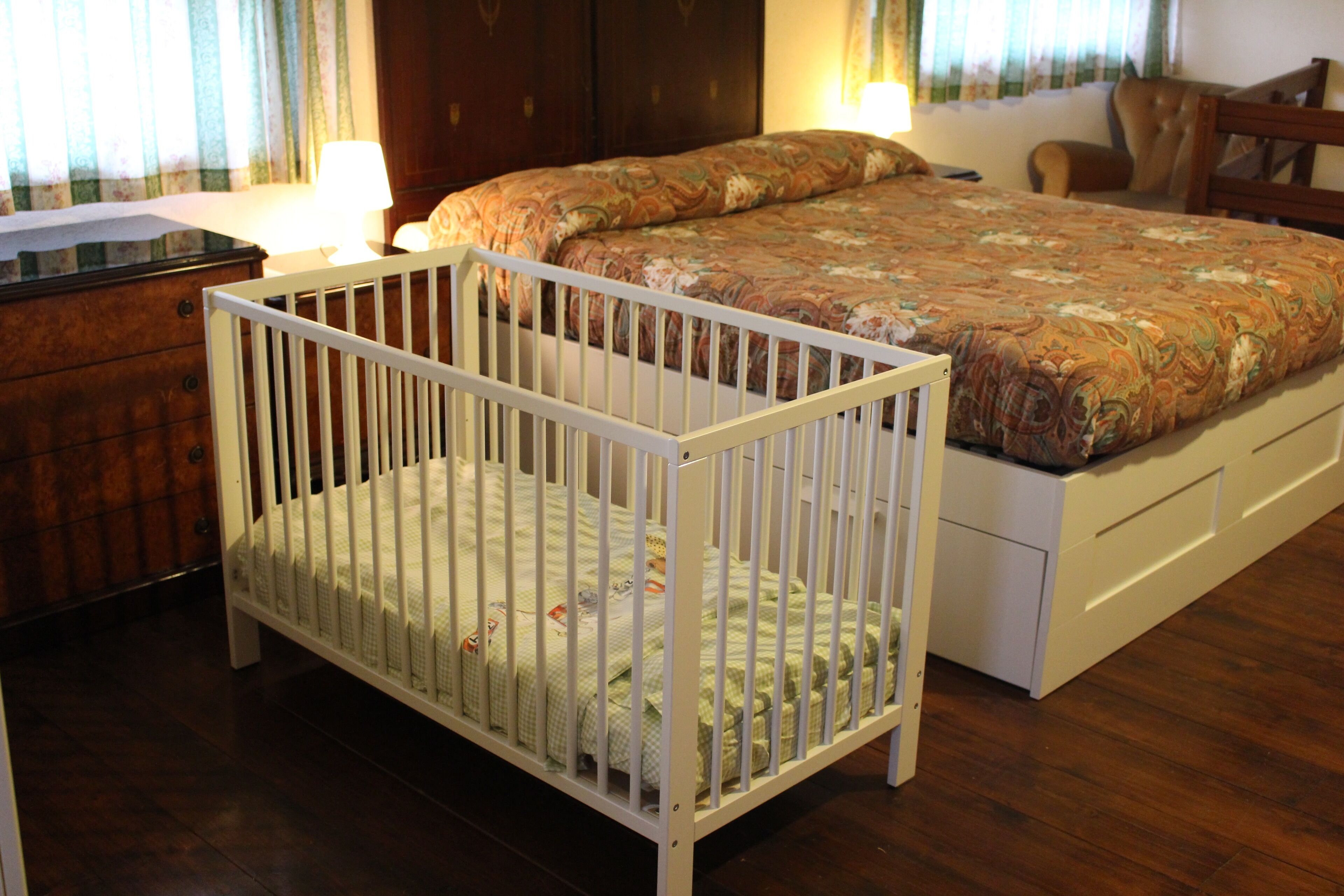 A room with a crib, a bed with a floral bedspread, a wooden dresser, and a chair.