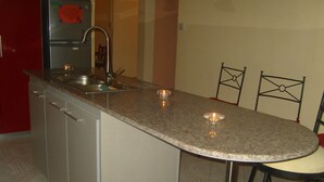 Une cuisine dotée d’un plan de travail en granit, d’un évier en acier inoxydable et d’un réfrigérateur. On y trouve deux chaises et une petite table.