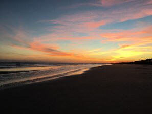 Ein Strand mit Sonnenuntergang, ruhiger See und Gebäuden in der Ferne.