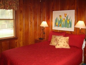 Ein Schlafzimmer mit einer holzverkleideten Wand, einer roten Bettdecke, zwei Lampen, einem Blumenbild über dem Bett und einem Fenster mit Jalousien.