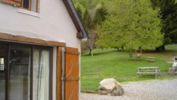 A rustic building with a large glass door and wooden shutters, set in a green landscape with trees and a bench. - Quiet chalet 5/7 places WIFI (Campan)