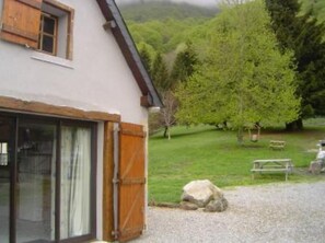 Un bâtiment rustique doté d’une grande porte vitrée et de volets en bois, situé dans un paysage verdoyant, agrémenté d’arbres et d’un banc.