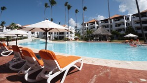 A resort pool area with lounge chairs and umbrellas, surrounded by palm trees and a building with multiple balconies. - Fantastic Condo on the Beach. Relax and enjoy the breeze !!! (Bavaro)