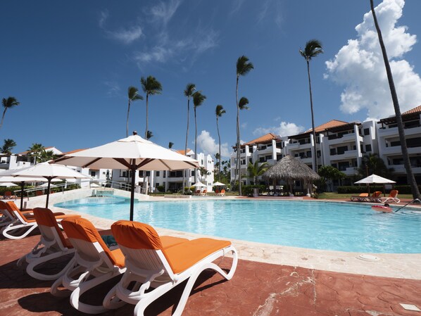 A resort pool area with lounge chairs and umbrellas, surrounded by palm trees and a building with multiple balconies. - Fantastic Condo on the Beach. Relax and enjoy the breeze !!! (Bavaro)