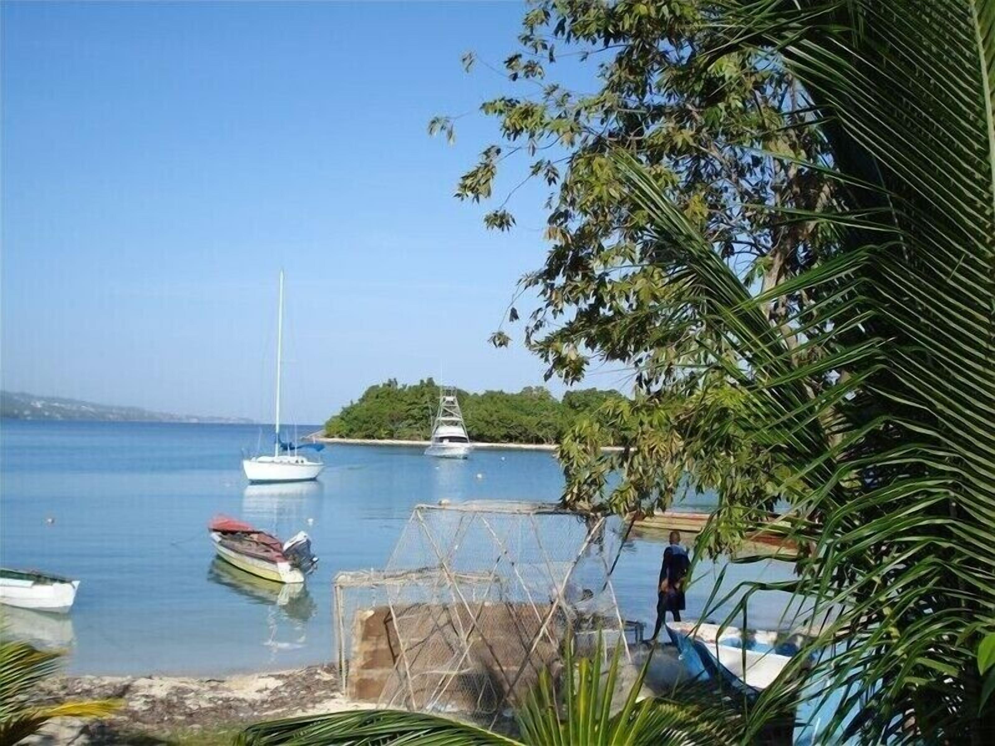 A calm bay with boats docked, a small island in the background, and a person walking along the shore.