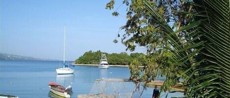 A calm bay with boats docked, a small island in the background, and a person walking along the shore.