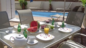 Zona de comedor junto a la piscina, con una mesa dispuesta para dos personas, platos, cubiertos y una variedad de frutas y bebidas.