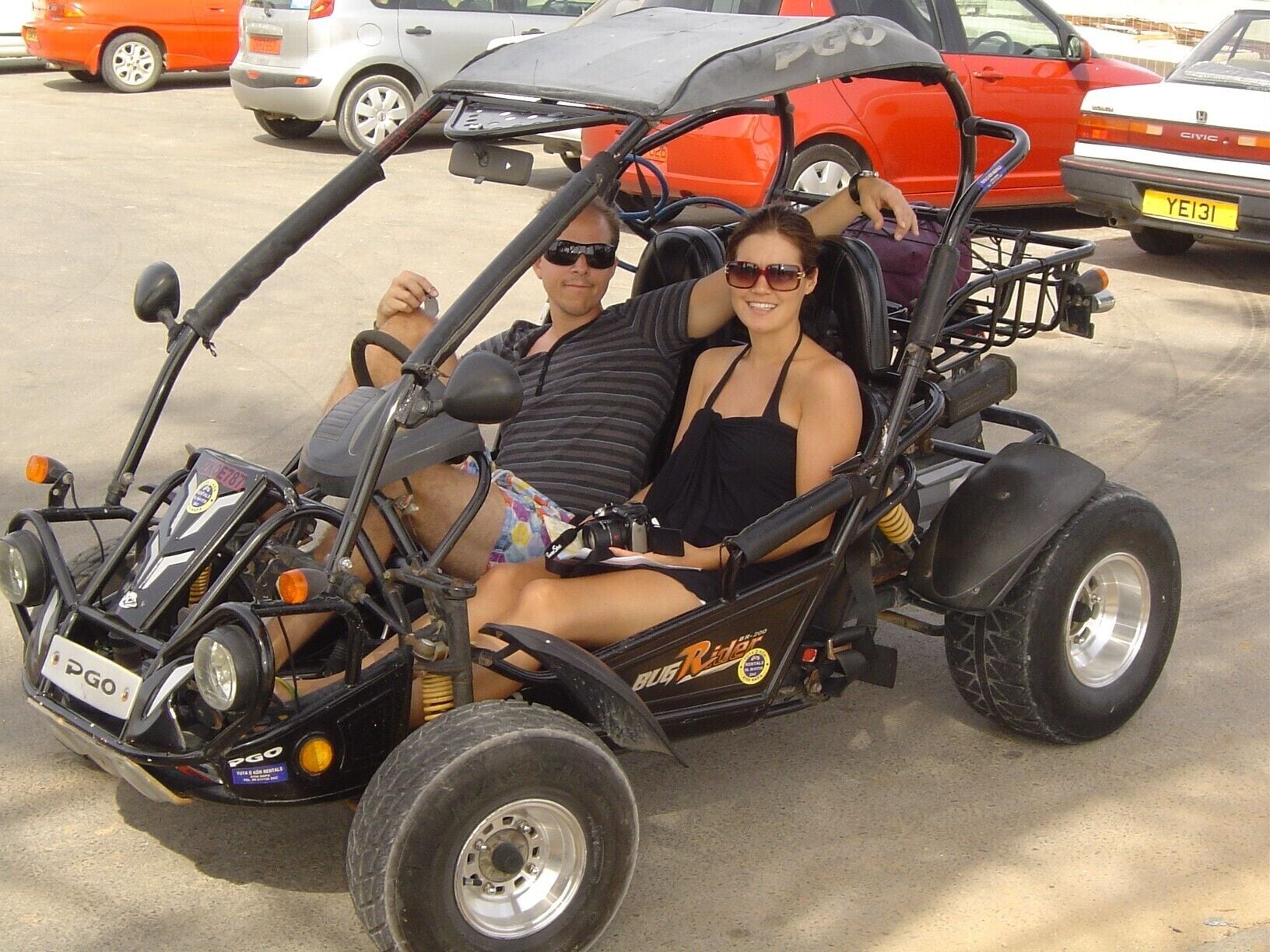 A couple in a small, open-top vehicle with a rugged design, parked in a lot with other vehicles.