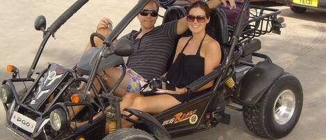 A couple in a small, open-top vehicle with a rugged design, parked in a lot with other vehicles.
