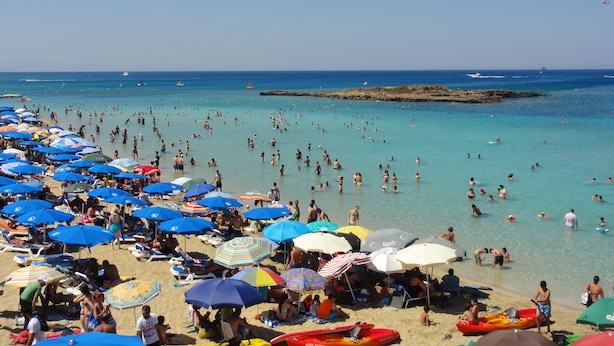 Una playa concurrida, con numerosos paraguas y gente disfrutando del agua.