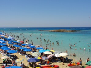 Una playa concurrida, con numerosos paraguas y gente disfrutando del agua.