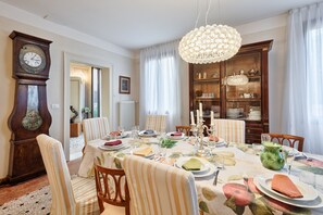 A dining room with a round table set for six, surrounded by six chairs. A large wooden bookcase is against the wall, and a grandfather clock is visible to the left.