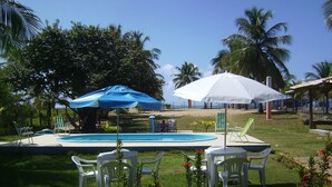 A pool area with white chairs and umbrellas, surrounded by greenery and palm trees. - Seafront house, paradise with privacy - ITACIMIRIM - 06 bedrooms and 4 suites (Camaçari)