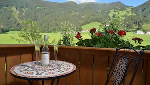 Een balkon met een betegelde tafel en stoelen, een fles wijn en uitzicht op glooiende groene heuvels en rode bloemen.
