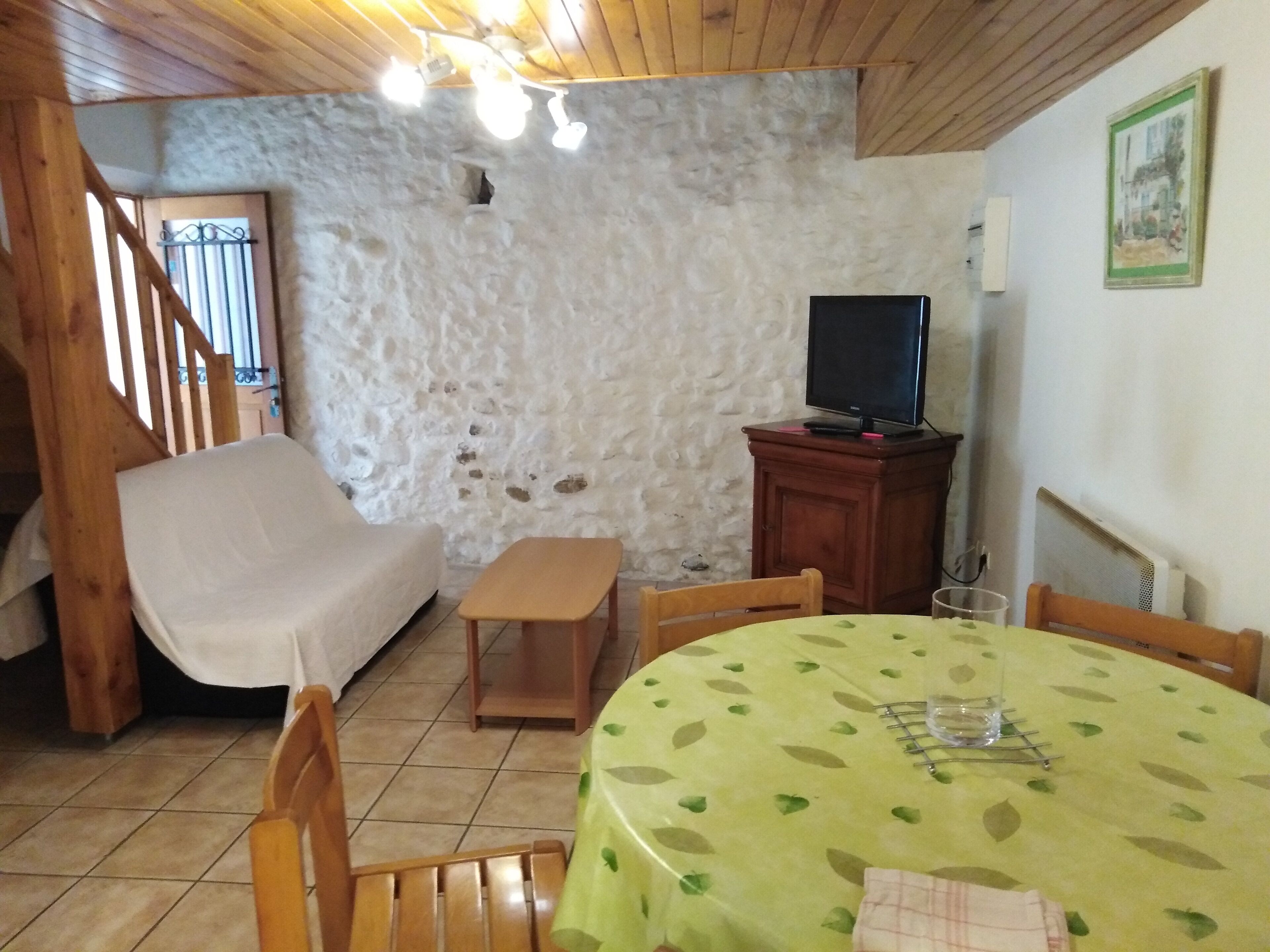 Une pièce chaleureuse avec un mur en pierre, une chaise en bois, une petite table recouverte d’une nappe à motifs de feuilles vertes, un téléviseur posé sur un meuble en bois et un escalier avec des rampes en bois.