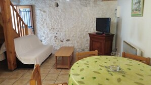 Una habitación acogedora con una pared de piedra, una silla de madera, una mesita con mantel de hojas verdes, un televisor sobre un mueble de madera y una escalera con barandillas de madera.
