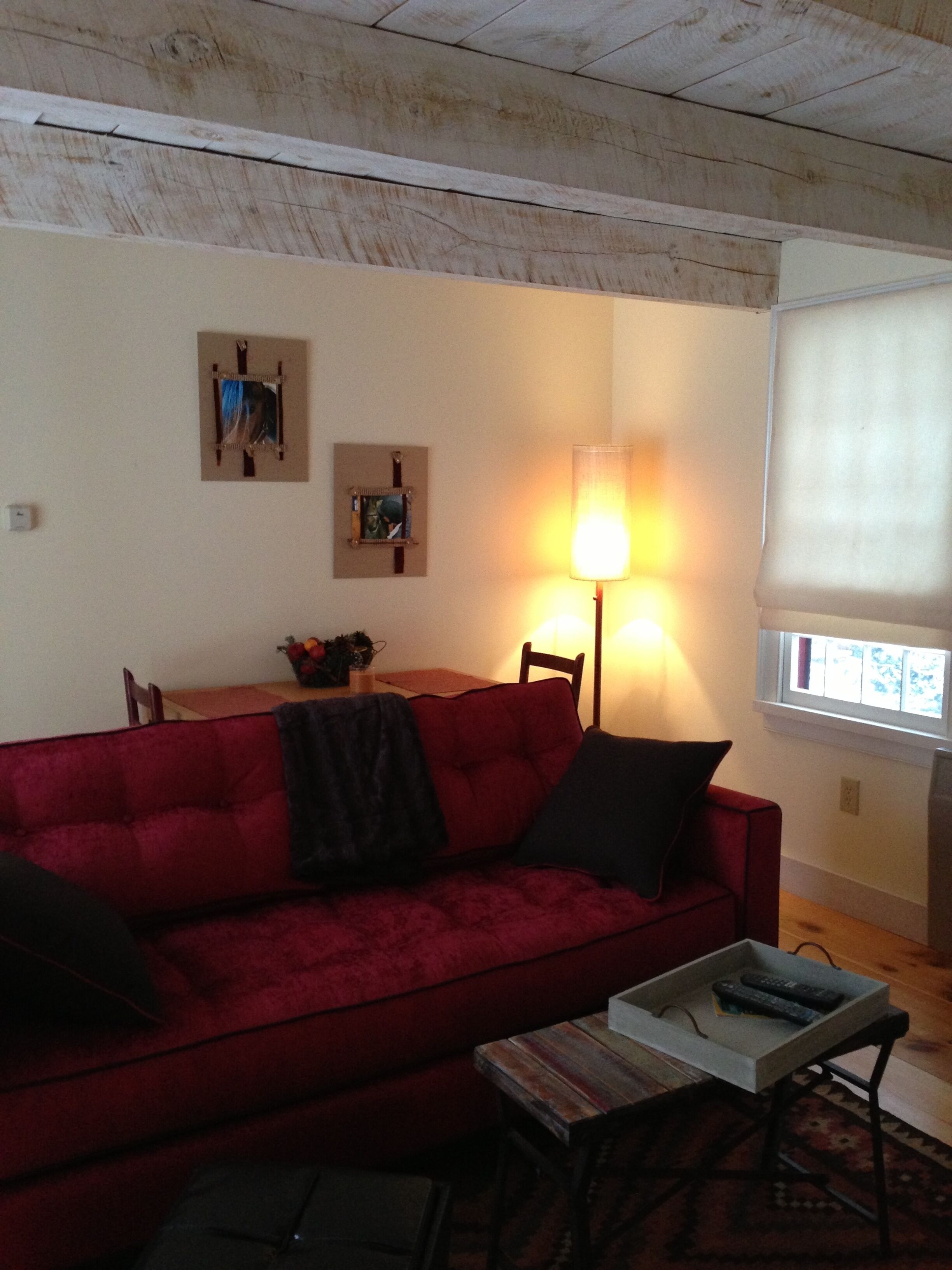 A living room with a red sofa, a coffee table, and a lamp. There is a window with blinds and a picture frame on the wall.