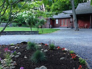 A rustic cabin with a stone foundation, surrounded by a gravel driveway and a landscaped garden with various plants and flowers. - Woodsy Country Cottage, HITS Horse Show, Fall Foliage, Skiing, Hunter Mt, Hiking (Saugerties)