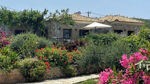 Un bâtiment en pierre, surmonté d’un toit en tuiles, entouré d’une végétation luxuriante et de fleurs aux couleurs vives.