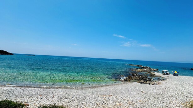 Une plage de galets bordée d’une eau turquoise et limpide, un rivage rocheux et quelques personnes abritées sous des parasols.