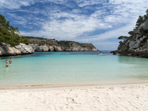 A beach with clear turquoise water, rocky cliffs, and a boat in the distance.