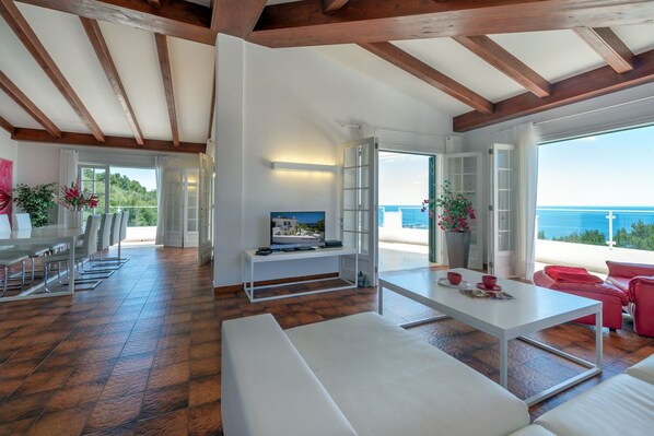 A spacious living area with a wooden ceiling, a white sofa, a dining table, and a TV.
