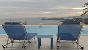 Two blue lounge chairs and a small table by a pool with a view of the sea and distant hills. - Villa Stella,  swimming pool, by the beach,  close to Athens
 (Marathon)