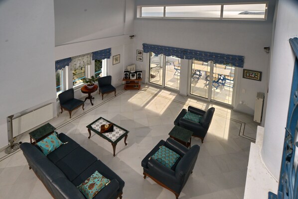 A spacious living area with a high ceiling, large windows, and a view of the outdoors. There are several sofas and chairs arranged around a central coffee table. - Villa Stella, swimming pool, by the beach, close to Athens
(Marathon)