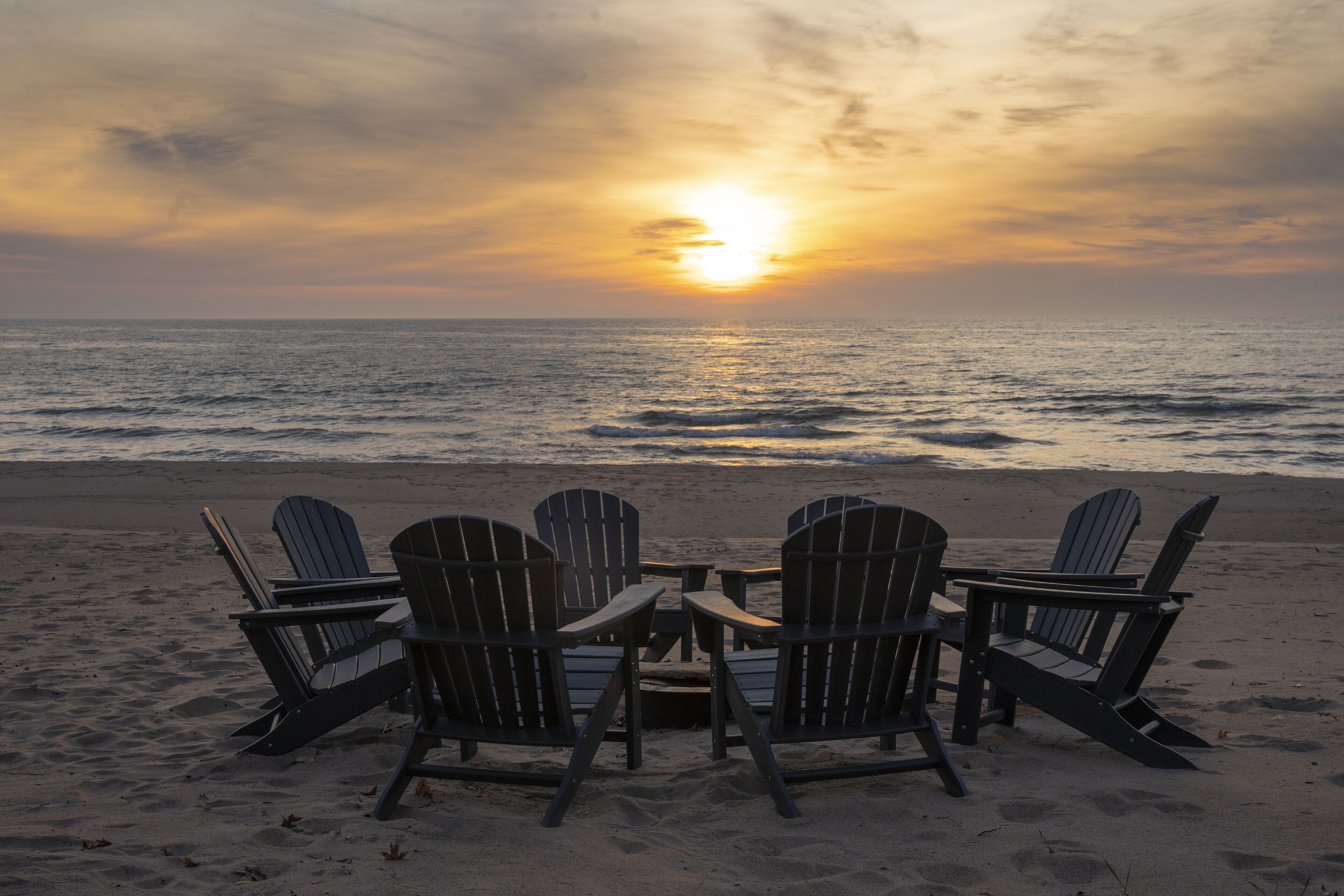 A beach scene with four Adirondack chairs facing the ocean at sunset.