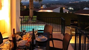Mesa de comedor dispuesta con platos, vasos y botellas, con vistas a una piscina y edificios a la distancia.