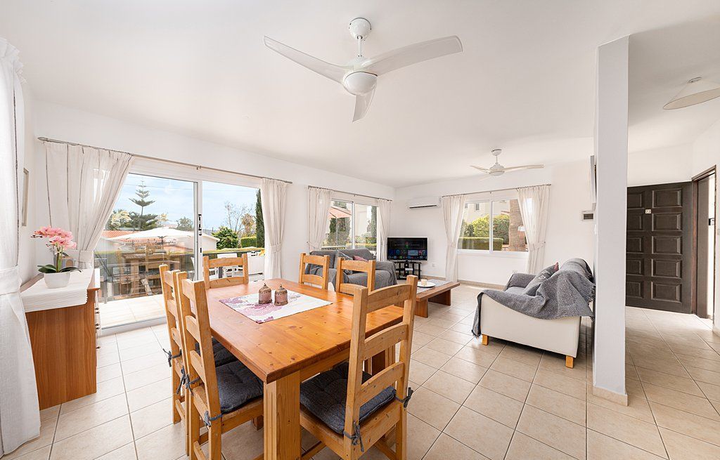 A spacious living area with a wooden dining table, a sofa, and a TV. There are large windows and a ceiling fan.