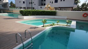 A swimming pool with a metal ladder, surrounded by a tiled deck and small palm trees.