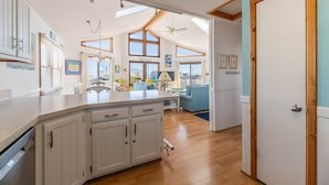 A spacious kitchen with wooden cabinets, a dining area with a blue sofa, and a view of the outside. - Key to Paradise - Kitty Hawk MP 4, Pool, Hot Tub, Excellent Ocean View, Easy Beach Access (Kitty Hawk)