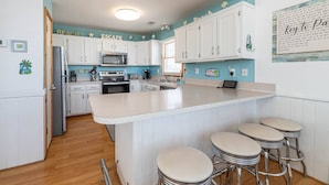 A kitchen with white cabinets, a central island with stools, a microwave, and a stove. - Key to Paradise - Kitty Hawk MP 4, Pool, Hot Tub, Excellent Ocean View, Easy Beach Access (Kitty Hawk)