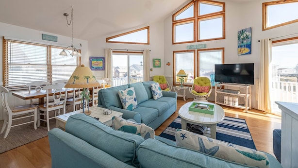 A spacious living area with a large sofa, dining table, and chairs. There is a television and a balcony with a view of the city. - Key to Paradise - Kitty Hawk MP 4, Pool, Hot Tub, Excellent Ocean View, Easy Beach Access (Kitty Hawk)