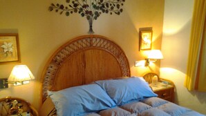 Une chambre d’hôtel avec une tête de lit en bois, un lit avec une literie bleue et beige, deux tables de chevet avec des lampes et des éléments de décoration murale, notamment un arrangement floral et des tableaux encadrés.