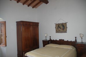 Une chambre à coucher avec une armoire en bois, un lit recouvert d’une couverture beige, deux tables de chevet avec des lampes et un tableau religieux accroché au mur.