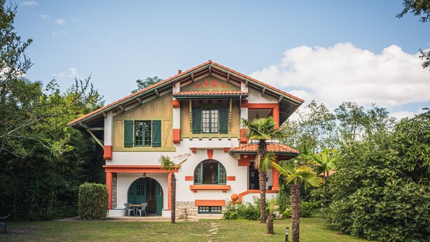 A two-story house with a red-tiled roof, green shutters, and a stone foundation.