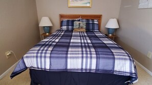 Un lit bien fait, recouvert d’une couverture à carreaux bleus et blancs, avec deux lampes de chevet et un tableau encadré au-dessus de la tête de lit.