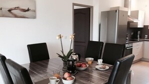 Uma sala de jantar moderna com uma mesa de madeira, cadeiras pretas e um vaso com flores. É possível ver uma cozinha ao fundo, com uma geladeira e armários.