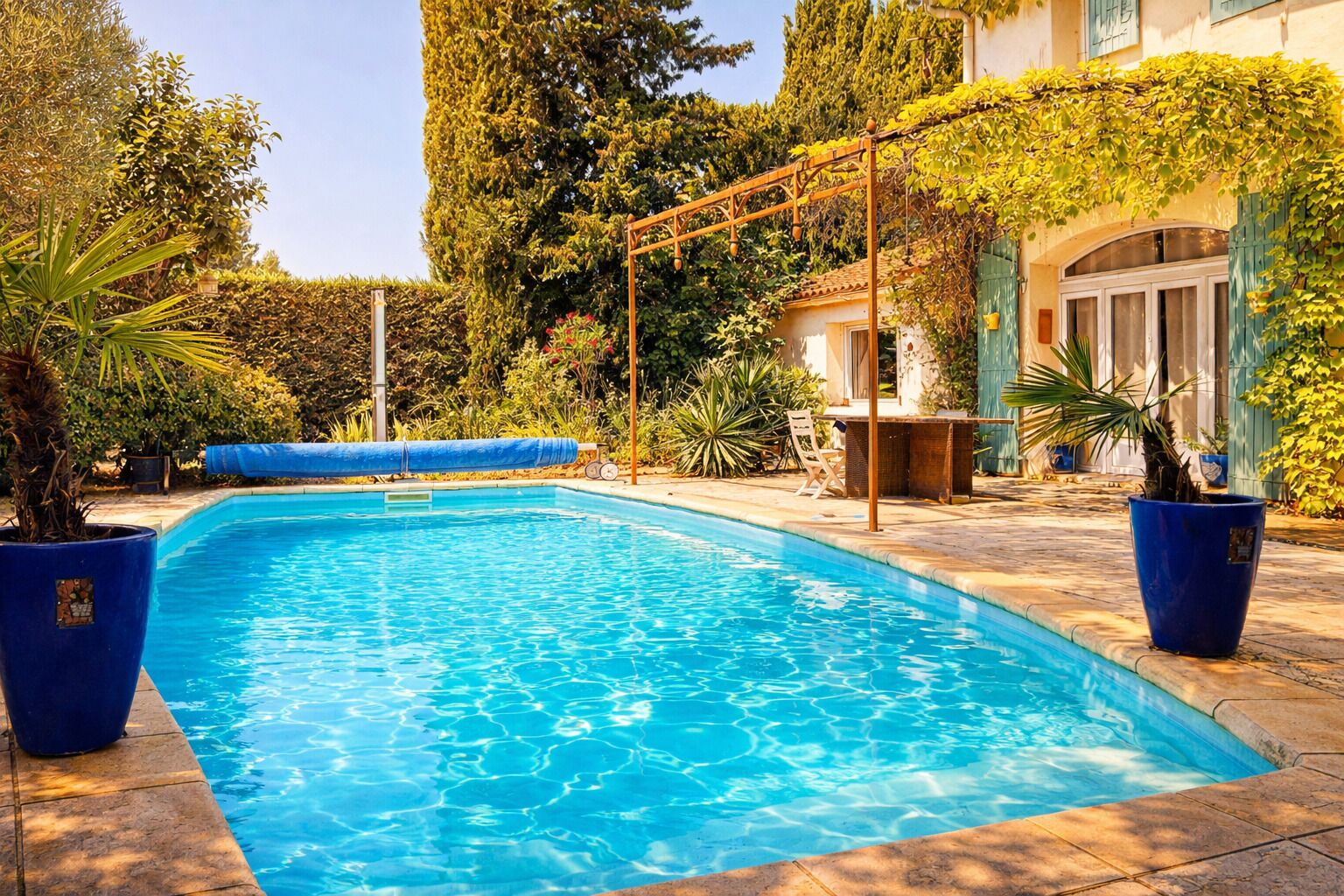 A swimming pool with a pergola, potted plants, and a building with a tiled roof in the background.