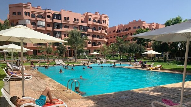 Ein Schwimmbecken mit Liegestühlen und Sonnenschirmen, umgeben von einem Gebäude mit mehreren Balkonen.