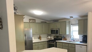 Una cocina moderna con armarios verde claro, un refrigerador de acero inoxidable, un microondas y un lavavajillas. Hay tres taburetes negros y una mesa de madera.