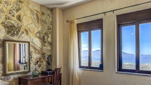 Ein Zimmer mit einer Steinsäule, einem Schreibtisch mit Stuhl, einem Bett und zwei Fenstern mit Blick auf das Meer und die Berge.