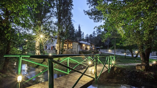 Un pont en bois enjambe un ruisseau et mène à une maison en pierre dotée d’une rambarde en métal vert.