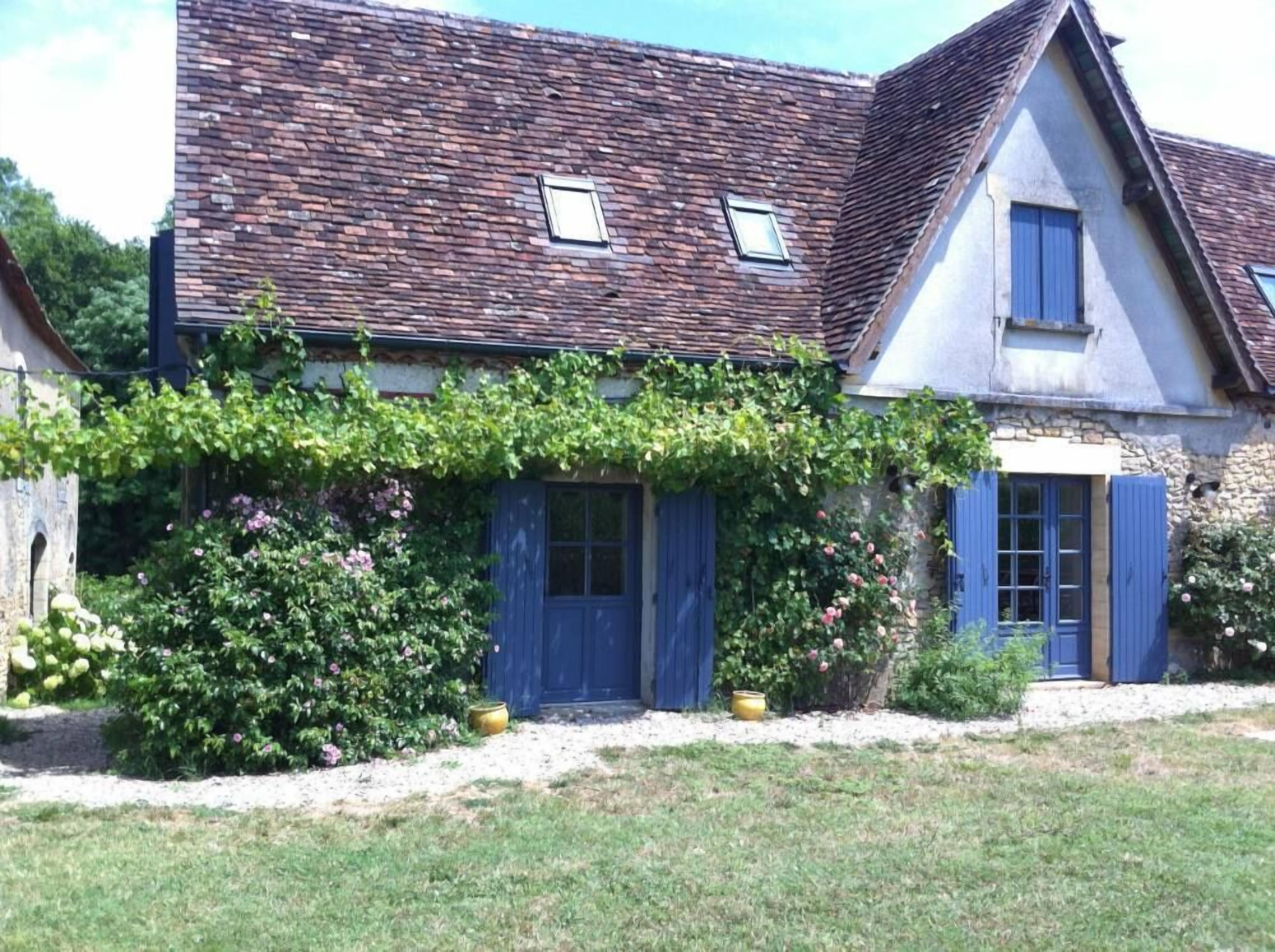 Ein traditionelles Steinhaus mit geneigtem Dach, blauen Fensterläden und einem Garten mit blühenden Pflanzen.
