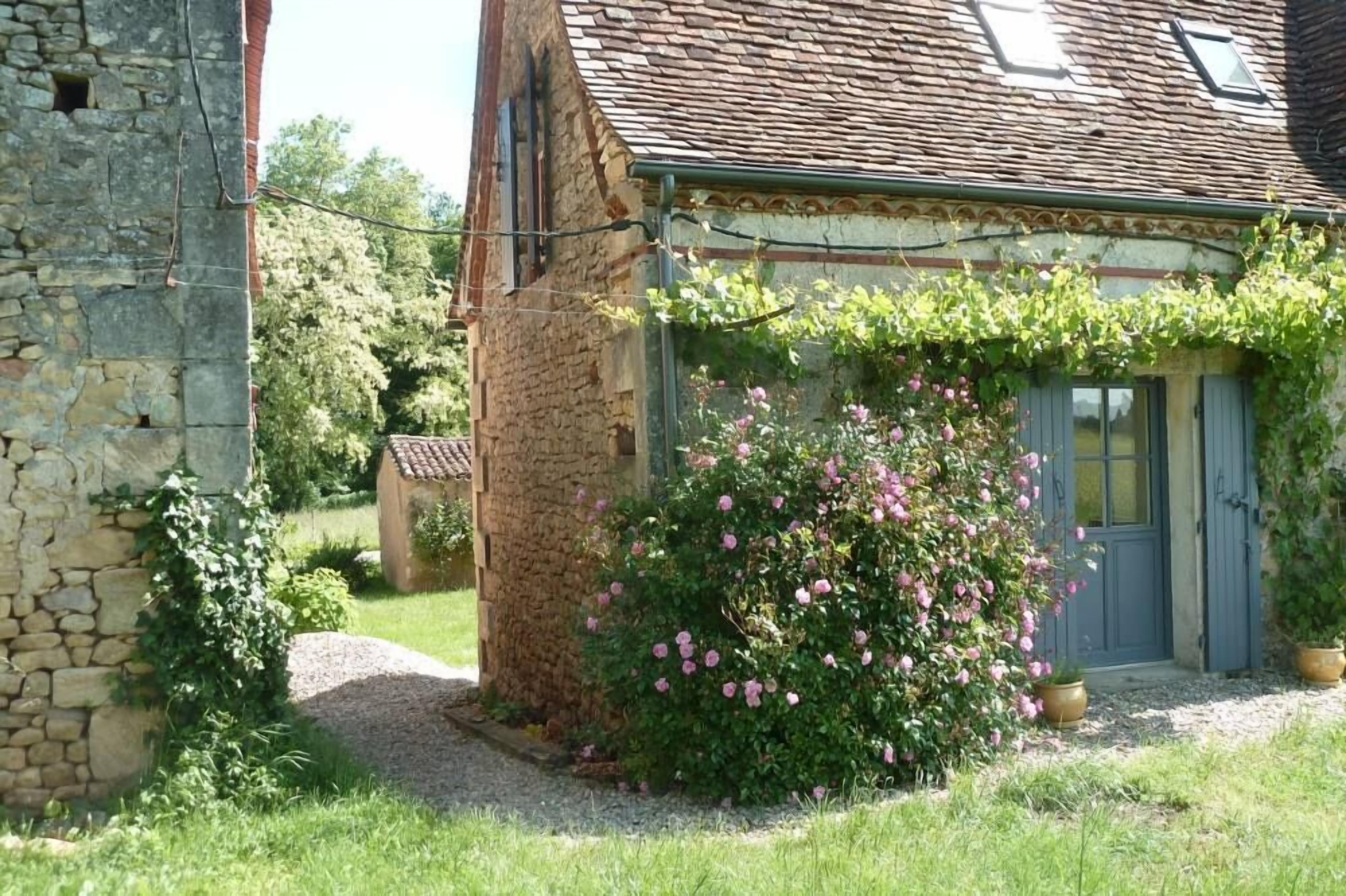 Un bâtiment rustique en pierre, doté d’un toit traditionnel recouvert de tuiles, d’un petit jardin agrémenté de fleurs roses et d’une porte avec des volets.