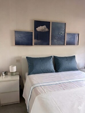 Hotelkamer met een bed, twee kussens, een nachtkastje met een lamp, een wandklok en een kunstwerk in een lijst boven het bed.