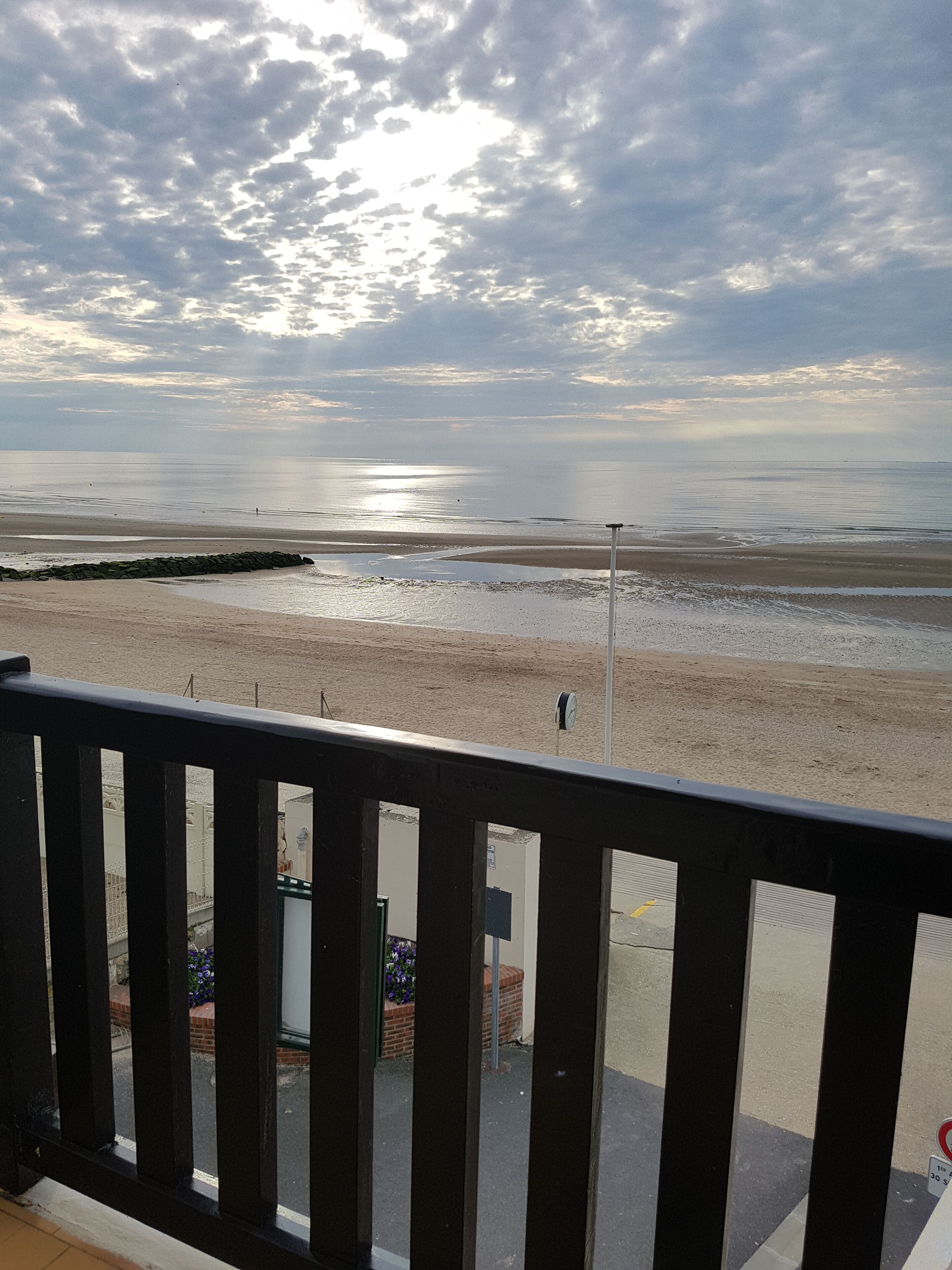 A beach view from a balcony with a railing, a street with buildings and a parked car, and a cloudy sky.